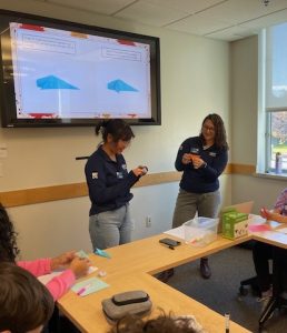 Two UConn tutors leading an origami lesson with a group of high school students. The current step in the origami lesson is displayed on a screen behind the tutors. Students are following along with a variety of brightly colored paper. 
