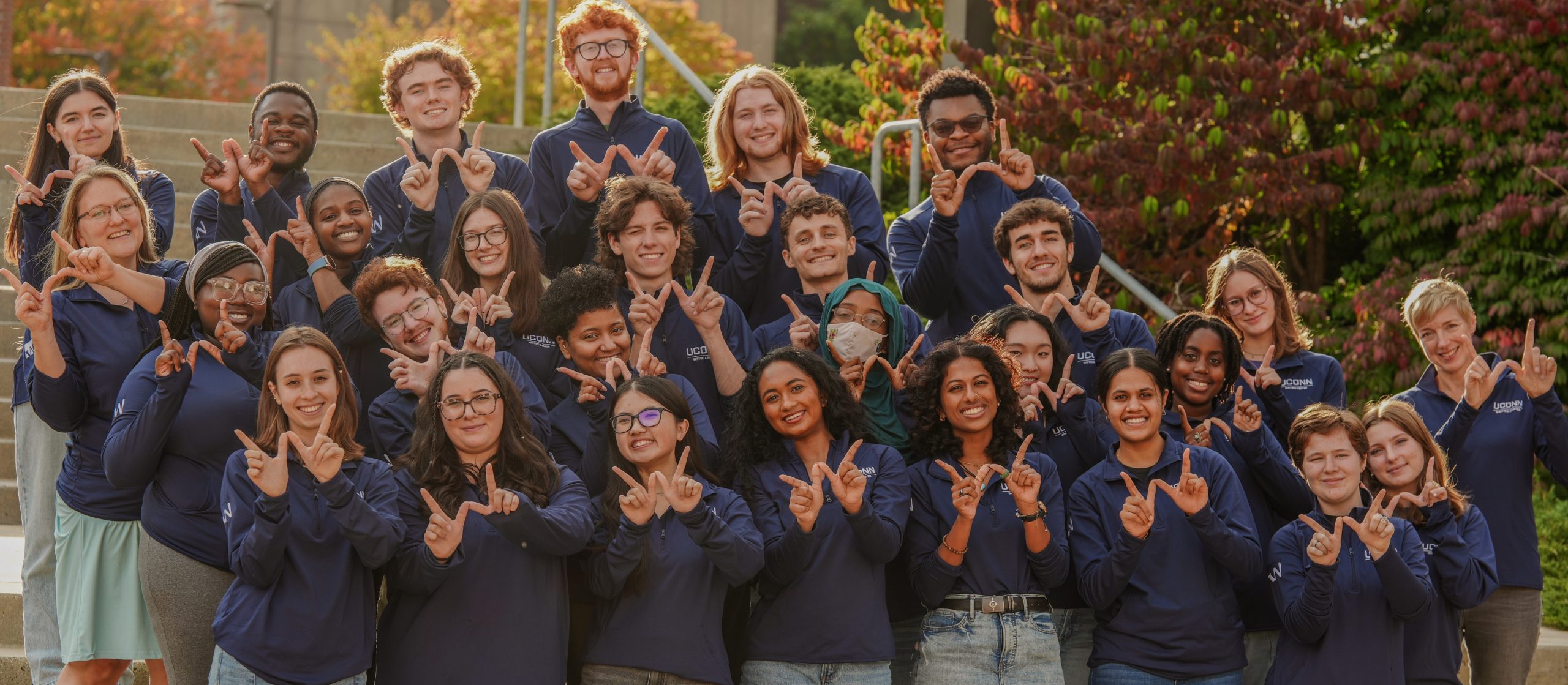 The members of the writing center staff posing outdoors on a set of stairs. Each person is holding up the "w" writing center logo and smiling.