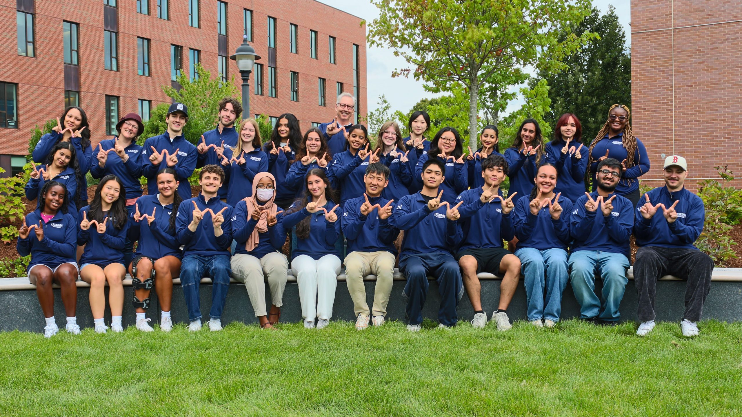 The members of the writing center staff posing outdoors. Each person is holding up the "w" writing center logo and smiling.